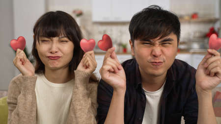 shoulder shot portrait of asian funny young couple holding paper heart over eyes and making funny faces while looking at the camera on home backgroundの写真素材