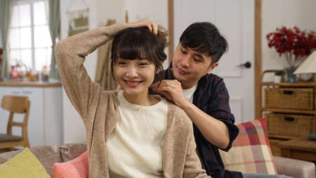 smiling asian woman holding up hair as her husband is putting on necklace for her, feeling happy about the beautiful gift. loving man hugging his wife on the sofa at homeの写真素材