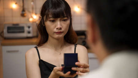over the shoulder shot of disappointed asian girlfriend getting unwanted present is closing the jewel box and politely smiling to her boyfriend on valentineâs day night date at homeの写真素材