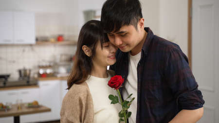 asian romantic couple holding a red rose and standing close together in a cozy home interior. the smiling wife is smelling the flower and hugging her husbandの写真素材