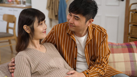 tilt up shot of a smiling pregnant asian woman looking and leaning on her husband on the sofa. the...