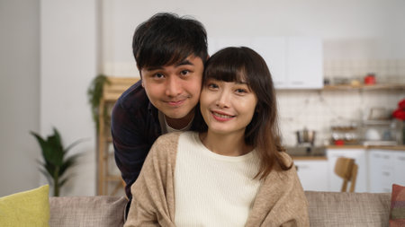 a loving asian man embracing his wife from behind while she is sitting on the sofa at home. they both look at the camera with a smileの写真素材