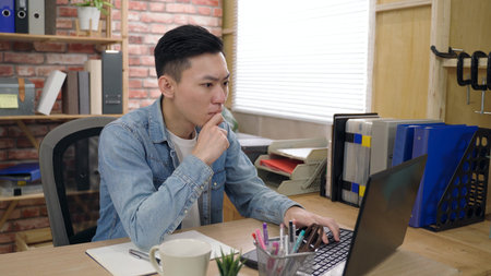 portrait asian male creative designer is taking note while working on the laptop with concentration...