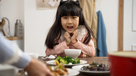 selective focus of amazed Asian daughter holding face with anticipation her mother is putting on delicious dish on dinner table at homeの写真素材