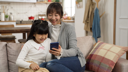 eisure asian little girl and her mom enjoying social media on smartphone at home. they cuddle up to...