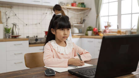 portrait of concentrated Japanese girl nodding head and taking notes while taking online class at home at dining table. her mother is busy cooking at backgroundの写真素材