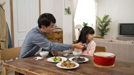 asian girl picky eater turning head and saying no to her dad as he is encouraging her to take a bite of vegetable. he holds the food with chopsticks near herの写真素材