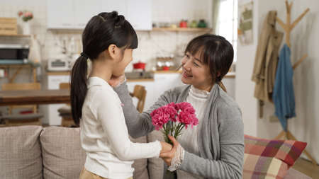 asian girl walking near to show surprise carnation bouquet to mother on mother's day at home. the happy mom smelling the flowers and smiling at her daughter while touching her faceの写真素材