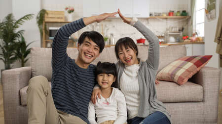 portrait of asian father and mother smiling at camera and making house roof figure with hands over their heads while sitting close with their daughter at homeの写真素材