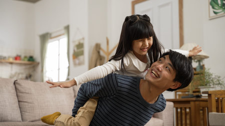 cheerful asian father carrying his cute daughter on back at home. the excited girl is pretending to...