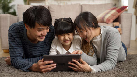 happy Asian family of three watching funny online video with tablet computer together at home. they lie prone on living room floor and point at screen while laughingの写真素材