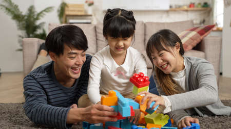 smiling Korean family of three having fun with building block together on ground at home. playful father and mother holding toys attacking each other beside the calm girlの写真素材