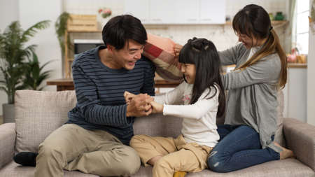 happy Asian Korean father mother and daughter playing tickle on living room couch at home. the woman having fun hitting the man with cushionの写真素材