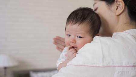 selective focus cute newborn infant is looking at camera innocently as her mommy is helping her burp at blurred background bright nursery room.の写真素材