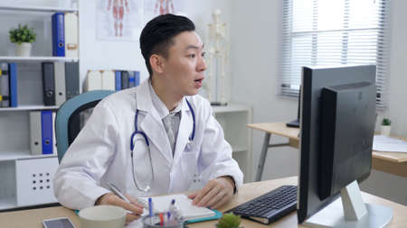 friendly family doctor sitting in front of the desktop is taking note while having an online consultation with his patient in the clinic.の写真素材