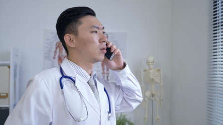 portrait asian male physician is receiving a phone call and talking with a serious look by the window in his office.の写真素材
