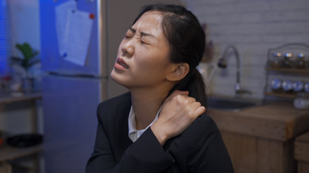 close up overworked asian businesswoman suffering from stiff neck is massaging her shoulders with painful expression in a home kitchen at midnight.の写真素材