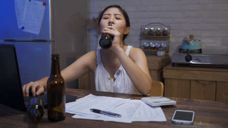 depressed asian wife worried about not having money to pay for debt is gulping beer and heaving a sigh while calculating daily expense on the laptop.の写真素材