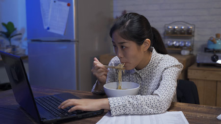 employee sitting down with midnight snack is stirring her noodles with chopsticks while typing...