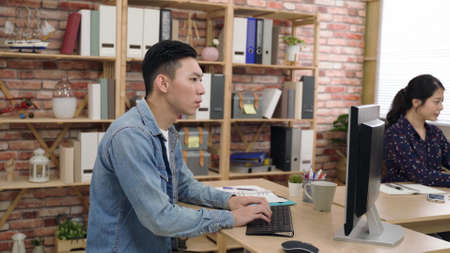 focused asian engineer is writing down data on his notebook with hand under chin while developing a computer program in an open creative office.の写真素材