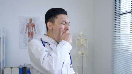 portrait tired resident doctor is yawning and exercising his stiff neck while having morning coffee by the window in his office.の写真素材
