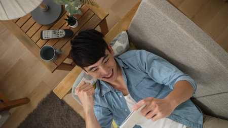 Top view cheerful Asian Japanese male lying on couch is celebrating with clenched fists for winning...