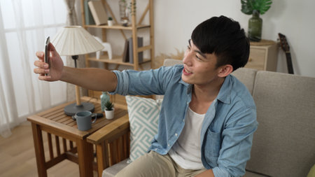 happy asian Korean male is waving hand and finger pointing to mobile phone screen while having fun chatting with friend through video call on the sofa at homeの写真素材