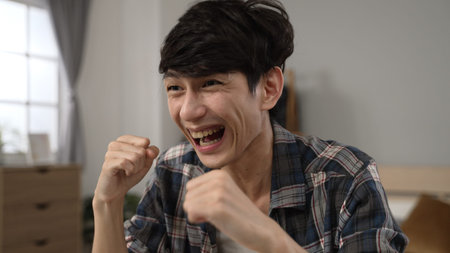 overjoyed asian japanese man cheering with clenched fists for online good news and covering mouth canât believe he got the great job offer on laptop at homeの写真素材