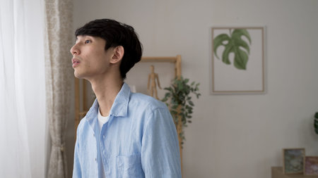 cheerful Asian young man leaving to welcome his friend after looking out the window waving hi with...