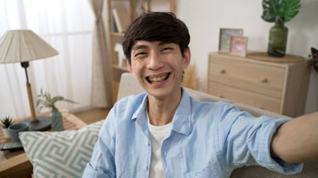 mobile cam view of cheerful asian Chinese guy waving hand and finger pointing at camera while having pleasant talk with friend via video call in the living room at homeの写真素材