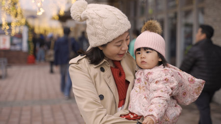 Japanese asian mother and her innocent baby in arms looking into distance while standing on a city street illuminated with Christmas lightsの写真素材