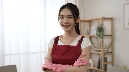 shoulder shot portrait of a pretty confident asian female in apron and gloves looking at the camera with a smile while posing at a modern living room backgroundの写真素材