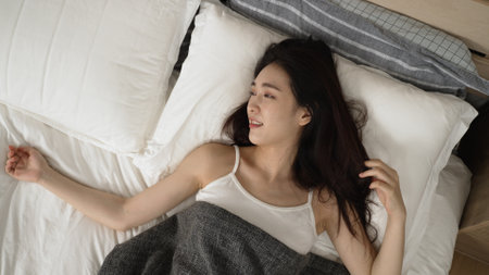 top angle of beautiful asian japanese woman just waking up on bed is touching hair and smiling with happiness while enjoying the tranquil morning in the bedroom at homeの写真素材