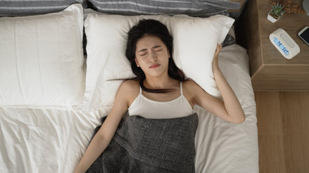 top angle view of tired asian chinese girl continue to sleep in after hand searching and hitting electric snooze alarm, unable to wake up in the morning on bed at homeの写真素材