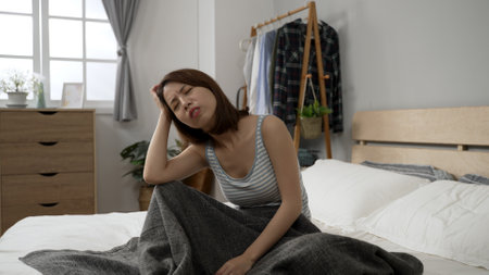 sad unhealthy young asian woman with migraine sitting on bed in modern bedroom at home. upset female with frowning face eyes and holding head with hand while suffering from strong sudden headache.の写真素材