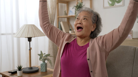 happy Asian retired old lady raising her arms while cheering for victory in mobile game she is playing on the phone in the living room at homeの写真素材