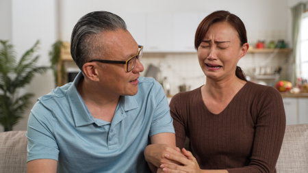 asian grandfather trying to comfort and encourage sad adult granddaughter on sofa at home. he taps her back while she covers her face feeling frustrationの写真素材