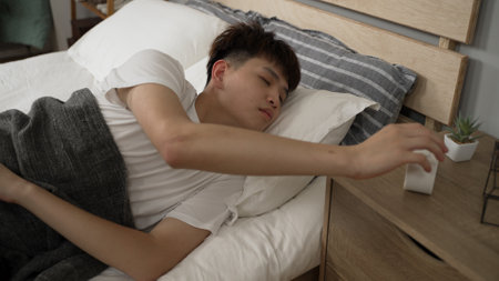 asian young man hitting the snooze button on the alarm clock and keep sleeping on bed in the morning in the bedroom at home.の写真素材