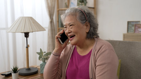 happy asian grandmother is smiling while enjoying chatting with friend on the mobile phone in a modern bright living room at homeの写真素材