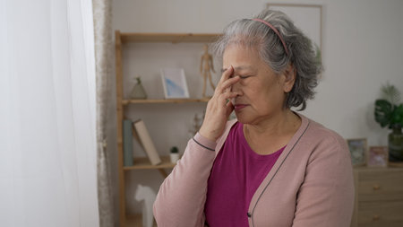 sad chinese old female is gazing out of window and rubbing her brows in contemplation while worrying...