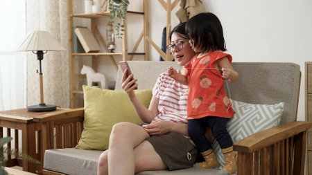 curious asian toddler baby girl playing with thread is looking at the mobile phone as her mother is scrolling on social media on sofa in the living room at homeの写真素材