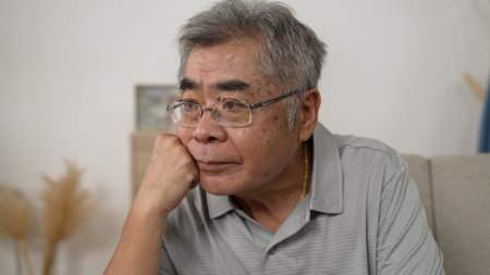 old age people and problem concept. senior Korean man in glasses thinking on sofa at home alone during virus outbreak. older korean male wearing gold chain and feel confused with hand on chin restの写真素材