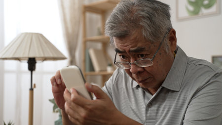 Elderly asian male having eyesight problems and wearing eyeglasses while gazing and reading message on smartphone on sofa. Old man having eye blurred vision indoors and moving glasses up and downの写真素材