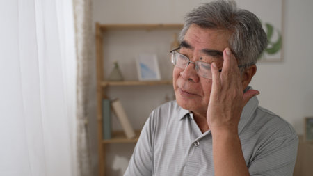 thoughtful middle aged Korean mature grandfather standing by window looking away. Pondering asian old senior hoary man think of psychological health problems spend time alone at homeの写真素材