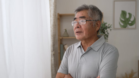 Side view thoughtful middle aged mature grandfather standing by window looking away. Pondering asian old senior hoary man think of psychological health problems spend time alone at homeの写真素材