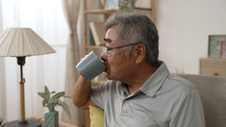 Waist up of senior man enjoy hot drink. elder male holding cup in hand sitting on sofa at home. old grandfather relaxing on couch with teaの写真素材