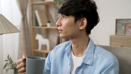 cheerful asian japanese young male looking out of window and enjoying the tranquil morning and coffee from cup in the living room at homeの写真素材