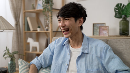 closeup of a cheerful asian Japanese male holding remote control is bursting out laughing while watching a comedy on tv in the living room at homeの写真素材