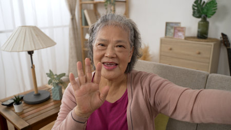 mobile cam view of a happy asian elderly woman waving hand and talking with a smile during a virtual chat with friend in the living room at homeの写真素材