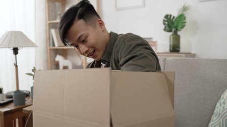 happy Korean male customer sitting down on couch to unpack the parcel carton box is surprised and satisfied with the giveaway package by postal shipping at homeの写真素材